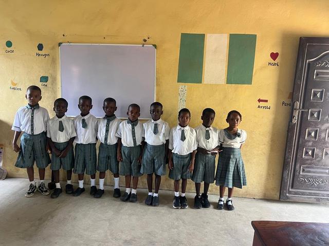 A class of pupils standing in front of the whiteboard and a painted Nigerian flag in their classroom